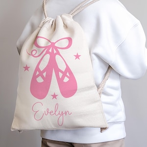 Può includere: Una borsa a sacco in tela bianca con ballerine rosa e stelle. La borsa ha il nome "Evelyn" stampato sul davanti.