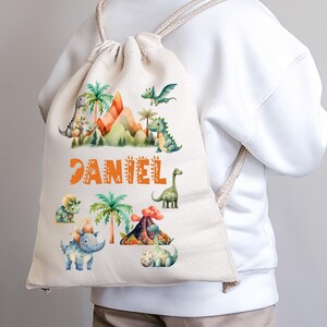 Może przedstawiać: Biały plecak z sznurkiem do ściągania w stylu dinozaurów. Plecak przedstawia kolorową ilustrację dinozaurów, gór i palm. Imię "DANIEL" jest nadrukowane pomarańczowymi literami na przodzie plecaka.