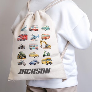 May include: A beige canvas drawstring backpack with a colourful print of various vehicles, including a bus, a helicopter, a tractor, and a motorcycle. The backpack has the name "JACKSON" printed on the front.
