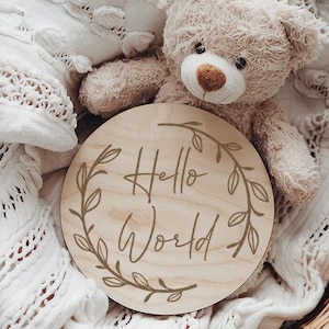 Op de afbeelding: Een rond houten bord met een krans van bladeren en de tekst "Hello World" in handschrift. Het bord ligt in een rieten mand met een wit gebreid deken en een beige teddybeer.