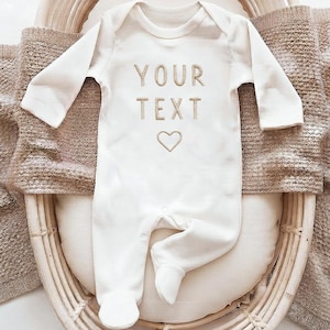 May include: A long-sleeved, cream-colored footed sleeper featuring the embroidered text "YOUR TEXT" in a tan font, positioned above a simple embroidered heart outline. The garment has a round neckline and integrated feet for full coverage.