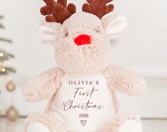 May include: A soft reindeer toy with brown antlers, a red nose, and a cream-coloured body. The text "Olivia's First Christmas 2026" is printed on the front, along with a heart symbol. A festive Christmas-themed plush toy.