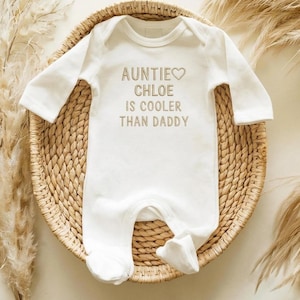 May include: White baby onesie with long sleeves and attached feet. The text "AUNTIE CHLOE IS COOLER THAN DADDY" is printed in a light brown color. The onesie is placed on a woven basket with dried plants in the background.
