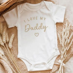 May include: A white baby bodysuit featuring the gold text "I LOVE MY Daddy" and a heart. The short-sleeved onesie is presented on a neutral blanket, with wheat stalks and a woven basket in the background.