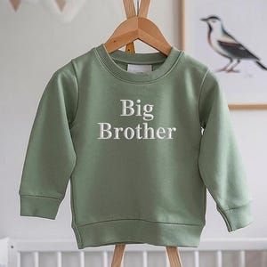 Op de afbeelding: Saliegroen sweatshirt met de woorden "Big Brother" in het wit geborduurd. De top met lange mouwen en ronde hals hangt aan een houten hanger. Een ingelijste vogelprint staat op de achtergrond.