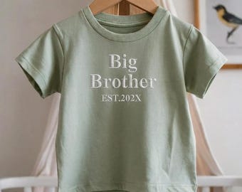 Może przedstawiać: Jasnozielona koszulka z białym haftem "Big Brother". Poniżej tekstu znajduje się "EST.202X". Koszulka jest prezentowana na drewnianym wieszaku.