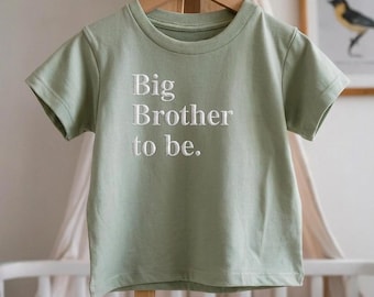 May include: Sage green t-shirt with the embroidered text "Big Brother to be." The short-sleeved shirt is displayed on a wooden hanger. The shirt is made of soft material and is suitable for a baby or toddler.