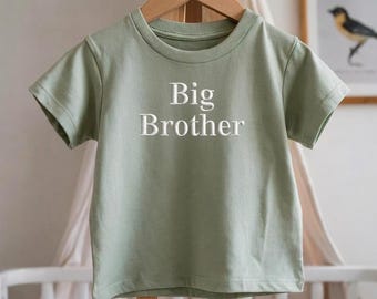 May include: A light green t-shirt with the words "Big Brother" in white block letters. The shirt is hanging on a wooden hanger. The shirt is short-sleeved and has a round neckline.