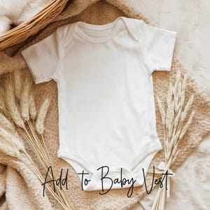 May include: A white baby bodysuit with short sleeves, lying on a soft, beige blanket. The text "Add to Baby Vest" is written in a cursive font at the bottom of the bodysuit. Dried wheat stalks are arranged around the bodysuit.