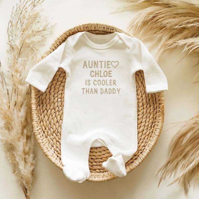 May include: White baby onesie with long sleeves and attached feet. The text "AUNTIE CHLOE IS COOLER THAN DADDY" is printed in a light brown color. The onesie is placed on a woven basket with dried plants in the background.