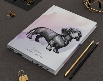 Puede incluir: Un diario blanco con un fondo de acuarela y una ilustración en blanco y negro de un dachshund. El diario dice "Dachshund My Dog Journal" en la portada.