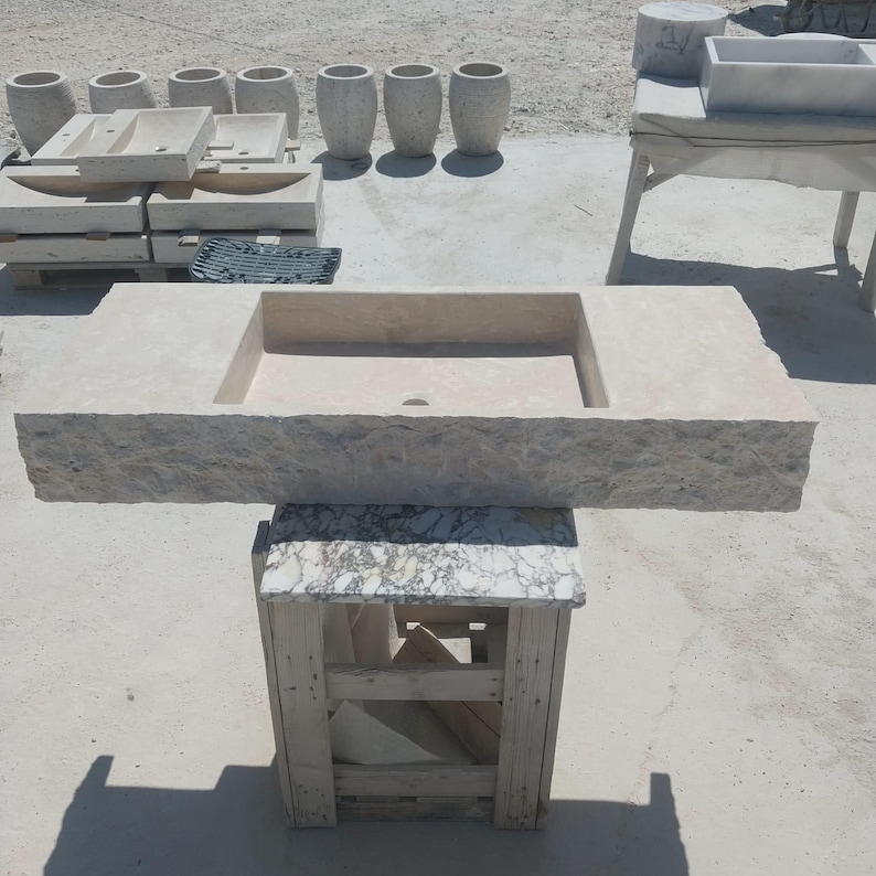 May include: A rectangular beige stone sink with a rough, chiseled front edge and a smooth basin surface. The sink is positioned on a wooden stand with a marble top. Several other stone sinks and cylindrical stone vessels are visible in the background.