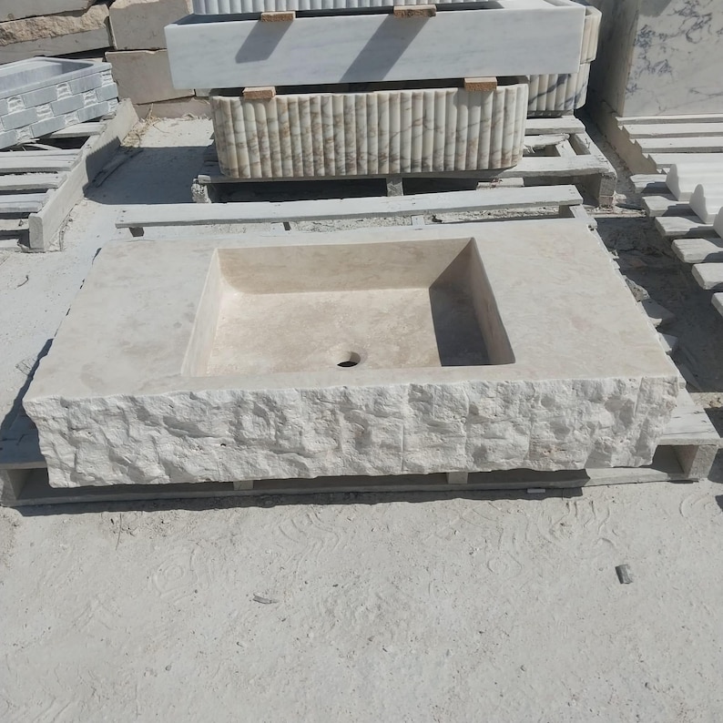May include: A rectangular beige stone sink featuring a smooth, flat basin area with a central drain hole and a textured, rough-hewn front edge. The sink is constructed from natural stone with visible variations in color and a matte finish.