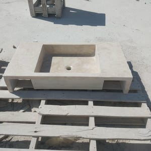 May include: A rectangular beige stone sink featuring a flat countertop surface with an integrated basin and a centered circular drain hole. The sink has a smooth, matte finish and is supported by two side legs, creating an open space underneath.