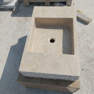 May include: A rectangular beige stone sink featuring a smooth interior basin with a central drain hole and a rough, chiseled exterior front edge. The basin is set into a flat, solid stone countertop surface with a natural, porous texture.
