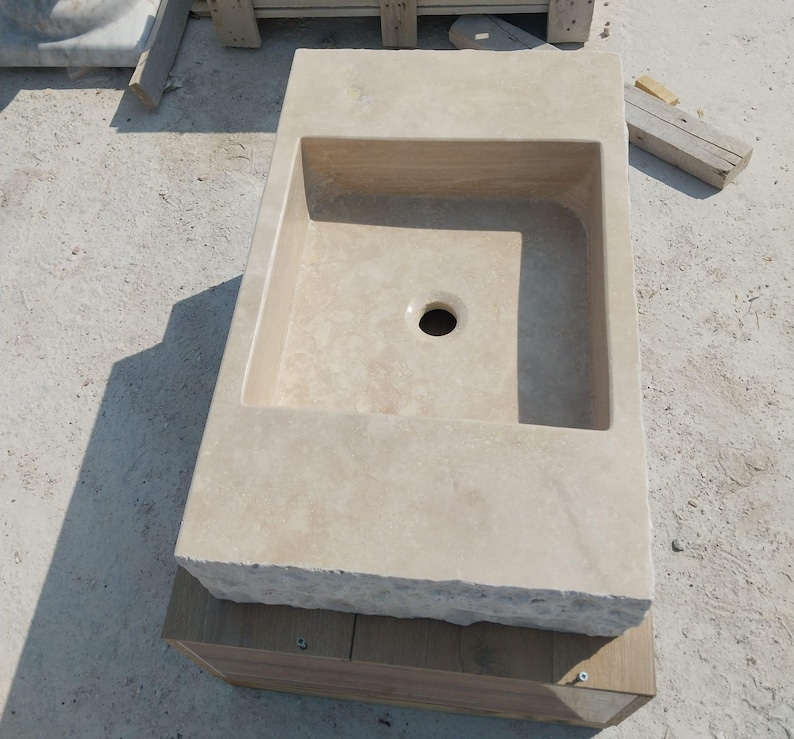 May include: A rectangular beige stone sink featuring a smooth interior basin with a central drain hole and a rough, chiseled exterior front edge. The basin is set into a flat, solid stone countertop surface with a natural, porous texture.