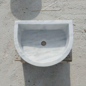 May include: A white marble sink with a semi-circular design. The sink has a smooth, polished surface with grey veining and a central drain hole. The sink is placed on a wooden support.