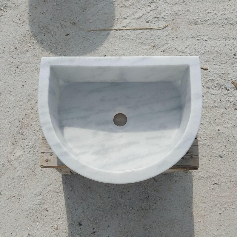 May include: A white marble sink with a semi-circular design. The sink has a smooth, polished surface with grey veining and a central drain hole. The sink is placed on a wooden support.