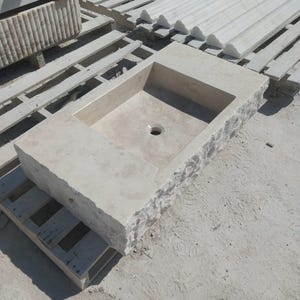 May include: A rectangular stone sink carved from light beige natural stone. The basin features a smooth, polished interior surface with a central drain hole, while the exterior sides retain a rough, chiseled, and textured rock-face finish.