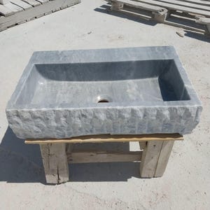 May include: A rectangular, gray stone sink with a textured exterior and a smooth interior basin. The sink features a central drain hole and is supported by a wooden pallet. The stone has a natural, weathered appearance, suitable for bathroom decor.
