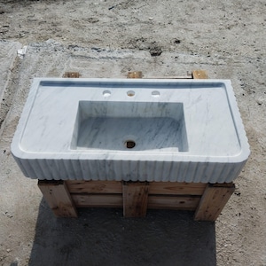 Può includere: Lavabo da bagno in marmo bianco con un bacino rettangolare e tre fori per i rubinetti. Il lavabo ha un bordo scanalato decorativo e si trova su una cassa di legno.