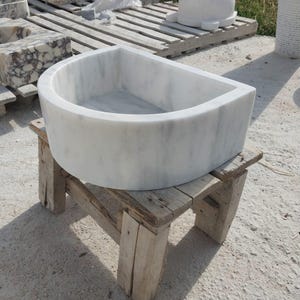May include: A white marble sink with a curved front and a rectangular basin. The sink is placed on a rustic wooden stand. The marble features subtle grey veining. This sink is designed for bathroom use and is made of natural stone.