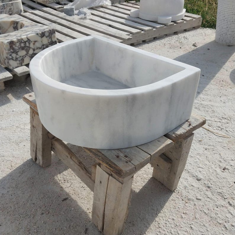 May include: A white marble sink with a curved front and a rectangular basin. The sink is placed on a rustic wooden stand. The marble features subtle grey veining. This sink is designed for bathroom use and is made of natural stone.