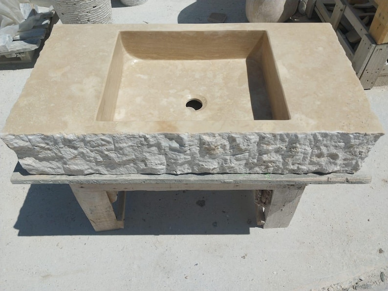 May include: A rectangular beige stone sink featuring a smooth, flat top surface with a central drain hole and a rough, chiseled, natural-looking exterior edge. The basin is deep and square-shaped, showcasing the natural texture and color variations of the stone material.
