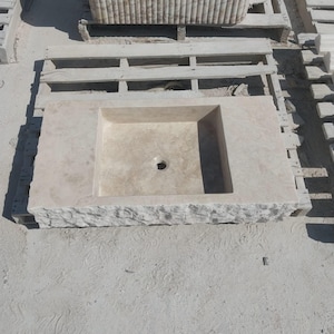 May include: A rectangular beige stone sink featuring a smooth, flat top surface with a central drain hole and a textured, rough-hewn front edge. The basin is recessed into the stone slab, showcasing natural variations in the light-colored stone material.
