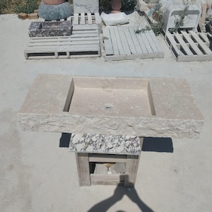 May include: A rectangular beige stone sink featuring a rough, chiseled front edge and a smooth, flat basin surface with a central drain hole. The sink is supported by a small, square pedestal base with a white and dark grey marbled stone finish.