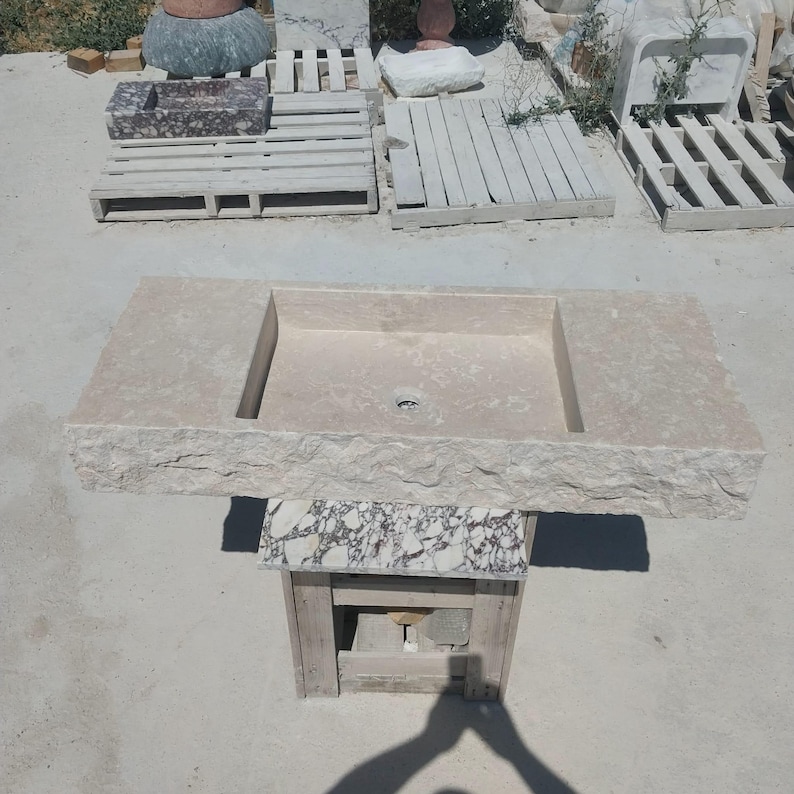 May include: A rectangular beige stone sink featuring a rough, chiseled front edge and a smooth, flat basin surface with a central drain hole. The sink is supported by a small, square pedestal base with a white and dark grey marbled stone finish.