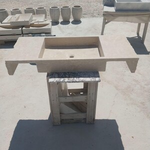 May include: A rectangular beige stone sink with a central drain hole and wide side ledges, supported by a wooden crate base. The sink features a natural, textured finish and is positioned outdoors among other similar stone basin components.