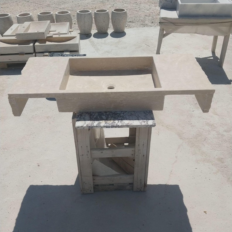 May include: A rectangular beige stone sink with a central drain hole and wide side ledges, supported by a wooden crate base. The sink features a natural, textured finish and is positioned outdoors among other similar stone basin components.