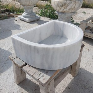 May include: A white marble sink with grey veining, semi-circular in shape, rests on a weathered wooden stand. The sink's smooth surface and rounded edges contrast with the rough texture of the stand. Other marble fixtures are visible in the background.