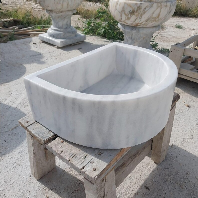 May include: A white marble sink with grey veining, semi-circular in shape, rests on a weathered wooden stand. The sink's smooth surface and rounded edges contrast with the rough texture of the stand. Other marble fixtures are visible in the background.