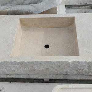 May include: A rectangular beige stone sink featuring a smooth, flat top surface with a central drain hole and a rough, chiseled, textured exterior edge. The basin is integrated into the solid stone block, showcasing natural variations in the light-colored stone.