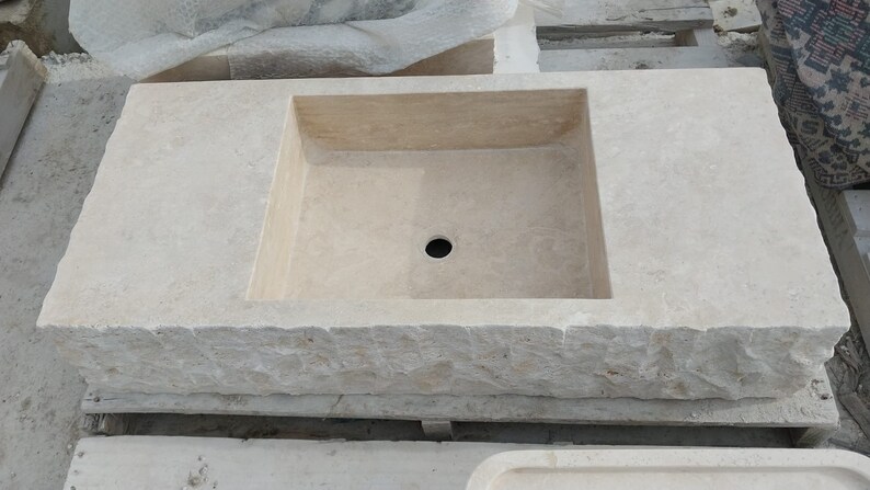 May include: A rectangular beige stone sink featuring a smooth, flat top surface with a central drain hole and a rough, chiseled, textured exterior edge. The basin is integrated into the solid stone block, showcasing natural variations in the light-colored stone.