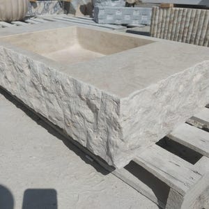 May include: A rectangular stone sink featuring a smooth, polished interior basin and a rugged, chiseled exterior texture. The natural beige stone displays subtle variations in color and grain, showcasing a raw, hand-hewn aesthetic on the sides.