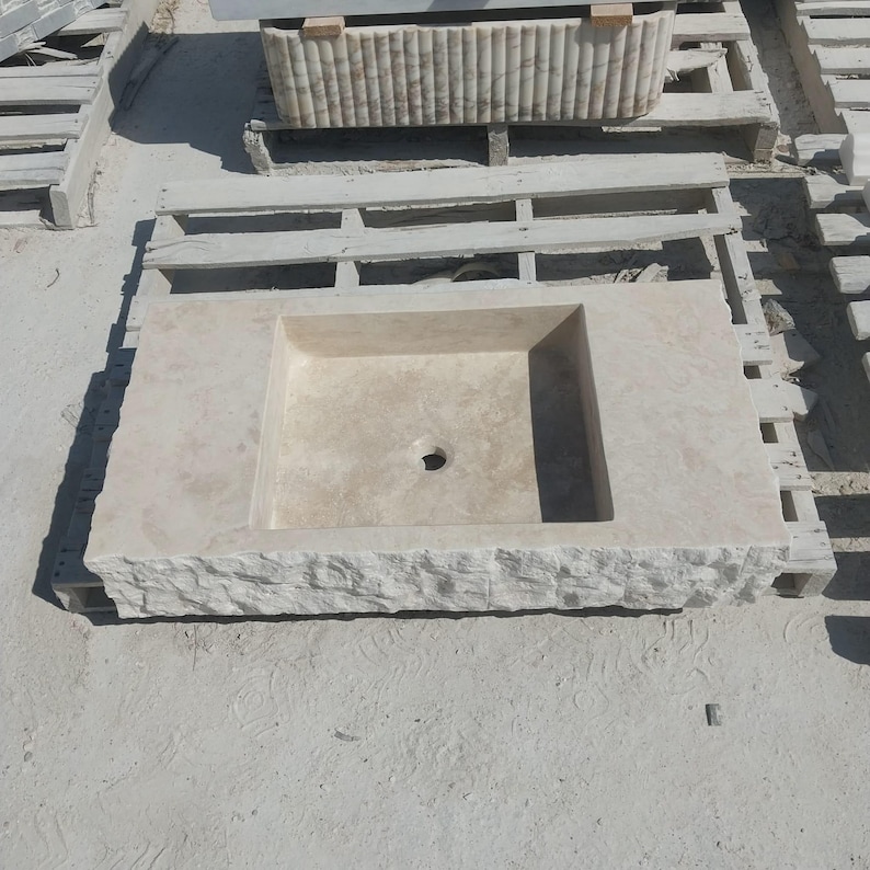 May include: A rectangular beige stone sink featuring a smooth, flat basin surface with a central drain hole and a rough, chiseled rock-face texture along the front edge. The sink is constructed from natural stone with visible light-colored veining.