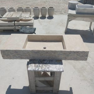May include: A rectangular beige stone sink featuring a rough, chiseled front edge and a smooth, flat top surface with a central drain hole. The sink is positioned on a wooden stand with a marble-patterned top, showcasing a natural, textured finish.