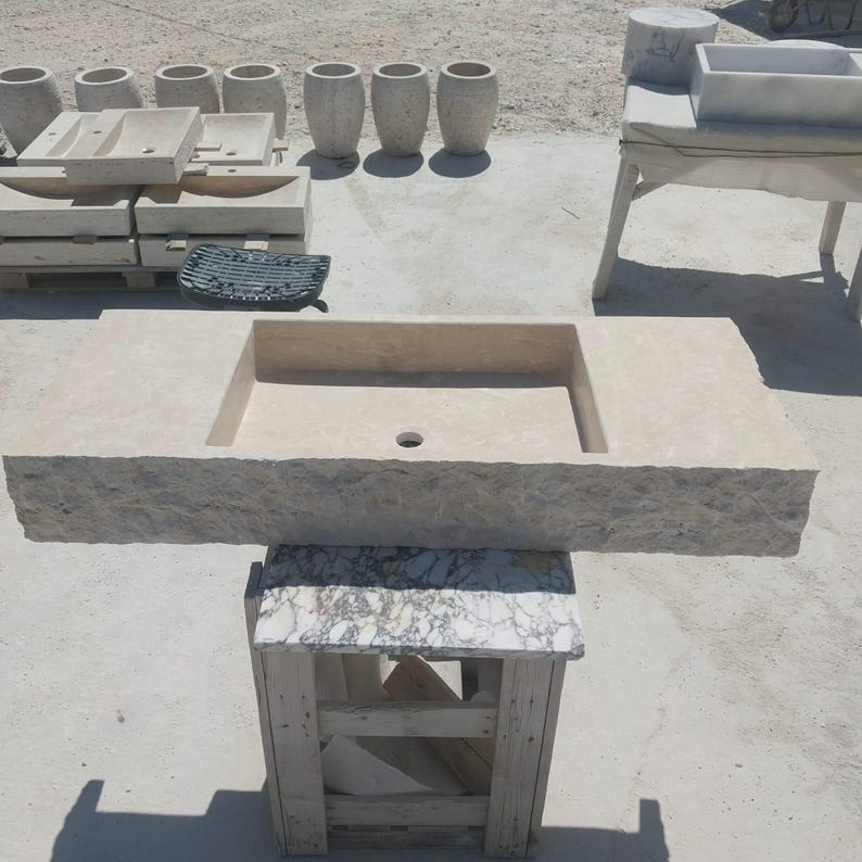 May include: A rectangular beige stone sink featuring a rough, chiseled front edge and a smooth, flat top surface with a central drain hole. The sink is positioned on a wooden stand with a marble-patterned top, showcasing a natural, textured finish.
