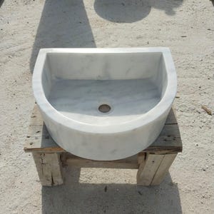 May include: A white marble sink with a semi-circular design. The sink has a smooth, polished surface and a drain hole in the centre. It is placed on a rustic wooden stand, showcasing its elegant simplicity.
