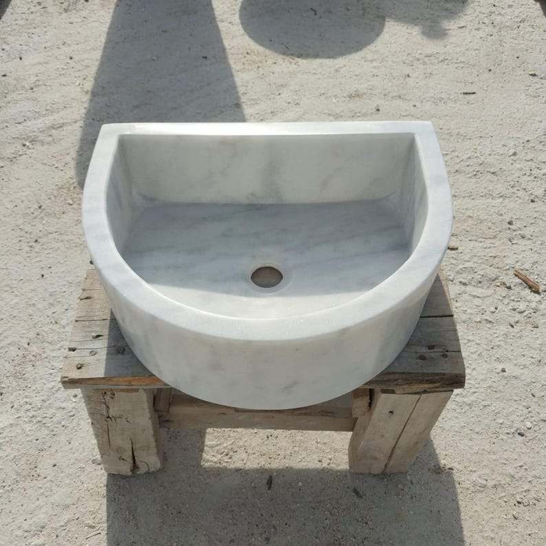 May include: A white marble sink with a semi-circular design. The sink has a smooth, polished surface and a drain hole in the centre. It is placed on a rustic wooden stand, showcasing its elegant simplicity.