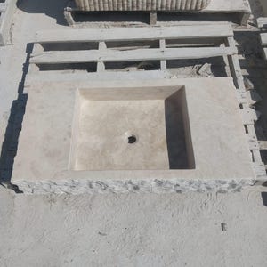 May include: A rectangular beige stone sink featuring a smooth, flat basin area with a central circular drain hole. The front edge of the sink displays a rough, chiseled, and textured natural stone finish, contrasting with the polished top surface.