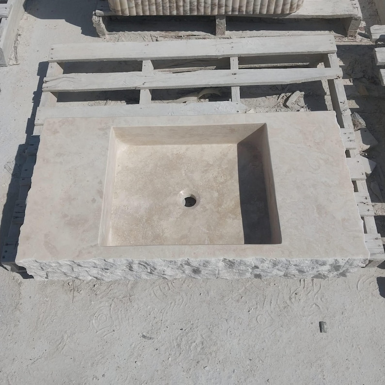 May include: A rectangular beige stone sink featuring a smooth, flat basin area with a central circular drain hole. The front edge of the sink displays a rough, chiseled, and textured natural stone finish, contrasting with the polished top surface.