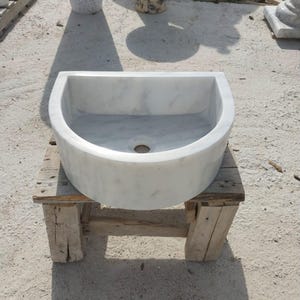 May include: A white marble sink with grey veining, semi-circular in shape, featuring a central drain hole. The sink is placed on a rustic wooden stand, showcasing its natural stone texture and design. The sink is a bathroom fixture.