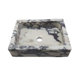 May include: A white marble sink with gray veining. The sink has a rectangular shape and a single drain hole in the center.