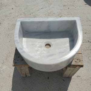 May include: A white marble sink with grey veining, semi-circular in shape. It has a central drain hole and sits on a rustic wooden stand. The polished surface reflects light, showing the natural stone patterns. A stylish bathroom fixture.