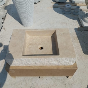 May include: A rectangular beige stone sink with a smooth basin and a central drain hole. The front edge of the sink features a rough, chiseled texture, while the top surface is flat. The sink is resting on a rectangular wooden support base.