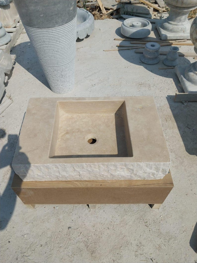 May include: A rectangular beige stone sink with a smooth basin and a central drain hole. The front edge of the sink features a rough, chiseled texture, while the top surface is flat. The sink is resting on a rectangular wooden support base.